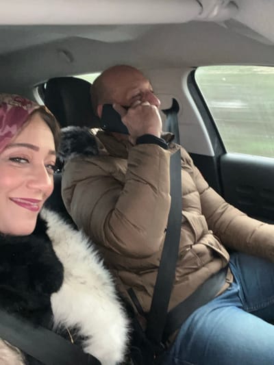 The image captures two individuals seated inside a vehicle, likely a car, during what appears to be an overcast day in Ain Johra, Morocco. The scene is presented from the perspective of a selfie being taken by the woman in the foreground.
The woman, positioned in the lower part of the frame, looks directly at the camera with a gentle smile. She has dark hair, dark eyes, and is wearing noticeable red lipstick. A patterned headscarf, featuring shades of maroon or dark red, is visible on her head. She is dressed in a black coat or jacket that features a prominent white, fluffy faux fur trim. A black seatbelt crosses diagonally across her.
Behind and to the right of the woman is a man, who is looking away from the camera, towards the front of the vehicle. He is wearing a beige or light brown puffer jacket with dark fur trim around the collar or hood. A black face mask covers his mouth and chin, and his left hand is raised to his nose or upper face, appearing to adjust the mask or touch his nose. He has short, light brown or bald hair. A seatbelt is also visible across his chest, and he appears to be wearing blue jeans.
Through the car window in the background, a blurry outdoor landscape suggests trees or foliage, with faint streaks on the glass possibly indicating rain or moisture. The light from outside is muted, suggesting an overcast sky. The image conveys a moment of travel, with one person taking a photo. No visible text is present in the image.