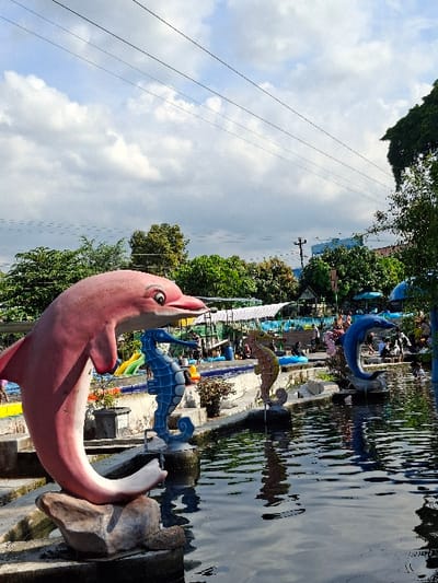 The image shows a pool with a pink dolphin statue in the foreground. The dolphin is facing right and its tail is in the water. Behind the dolphin is a blue seahorse statue. There is a second seahorse statue in the background, facing the dolphin. A third, blue seahorse statue is in the water behind the dolphin. The pool is surrounded by a concrete edge. The background features a green lawn, trees, and a blue sky with white clouds. People are visible in the background. The image was taken at a location in Kecamatan Tulung, Indonesia, at coordinates -7.592, 110.65. There are power lines running across the top of the image.