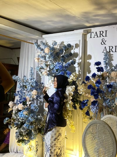 In Kembang Harum, Indonesia, a woman wearing a navy blue hijab and a matching satin jacket is posing for a photo in front of a decorative backdrop at what appears to be an event. The backdrop features an arrangement of artificial flowers in shades of blue, white, and peach, with some yellow floral accents hanging down. A white curtain hangs behind the floral arrangement. Text reading "TARI & ARI" is visible on the wall to the right of the backdrop. Two white chairs with woven seats are positioned to the right of the frame. To the left of the frame, a person in a purple shirt is partially visible, bending down. The lighting is warm and appears to be artificial, creating shadows on the wall. The ceiling shows exposed beams.