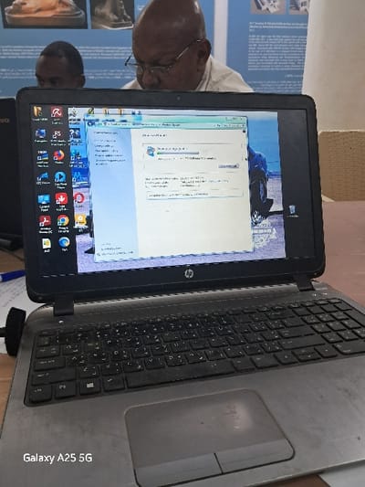 A silver HP laptop computer with a black keyboard is open on a wooden desk, with its screen displaying a progress bar that is 75% complete, indicating a download or update in progress. The laptop is in a room with a white wall and a light blue ceiling, with a person with black hair partially visible at the lower left edge of the image. The person appears to be looking at the laptop screen.  The laptop is the primary subject of the image, with the person in the background. The laptop screen displays an open window with a title bar that includes "Central Point Office." The laptop appears to be in the town of Karima in Sudan, located at coordinates 18.54 degrees latitude and 31.831 degrees longitude. The scene suggests a professional office environment.