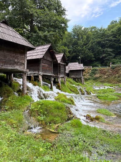 This media file depicts a picturesque scene in Ćusine, Bosnia and Herzegovina, featuring a series of traditional wooden watermills situated alongside a cascading waterfall. The watermills are constructed with rustic wooden planks and feature angled roofs, with several of them visible in a line along the hillside. They appear to be part of an operational setup, with the water from the falls flowing beneath and around them.
The surrounding environment is lush and green, with abundant vegetation and trees forming a dense backdrop. The waterfall itself is a prominent element, characterized by white, frothy water tumbling over moss-covered rocks and greenery. The water appears clear and energetic, suggesting a natural and unspoiled setting.
In the background, a person can be seen standing near the top of the waterfall, adding a sense of scale to the scene. The sky is partly cloudy, with patches of blue visible, indicating fair weather. The lighting suggests it is daytime, likely mid-morning or afternoon, with soft natural light illuminating the scene. The overall impression is one of tranquility and natural beauty, with the man-made watermills harmoniously integrated into the landscape. No visible text is present in the image.