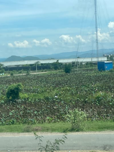 The image captures a vast expanse of a lotus field under a partially cloudy sky. In the foreground, a paved road runs horizontally, with a hint of vegetation on its edge. The lotus field dominates the mid-ground, filled with lush green lily pads and scattered pink and white lotus flowers. Beyond the field, a row of greenhouses with white covers stands tall, interspersed with smaller structures and trees. To the right, a tall communication tower pierces the sky, accompanied by a small, bright blue building at its base. In the distance, a series of rolling hills form a picturesque backdrop against the horizon. This scene is located in the city of Hồng Sơn, Vietnam.