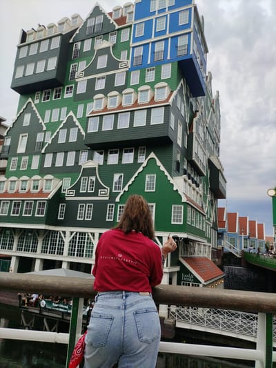 In Zaandam, Netherlands, a person wearing a red t-shirt and blue jeans stands on a wooden bridge, facing away from the camera, and looking at the Inntel Hotels Amsterdam Zaandam. The hotel is a modern, multi-story building with a distinctive stacked architectural style, resembling a series of traditional Dutch houses. The facade is a mosaic of colors, primarily green and blue, with white-framed windows and red-tiled roofs. The building's design creates an illusion of stacked homes. Below the hotel, there is a canal with a walkway and a sign for "Food & Drinks." Other buildings with similar Dutch architectural elements are visible in the background, along with a bridge and people milling about. The sky is overcast.