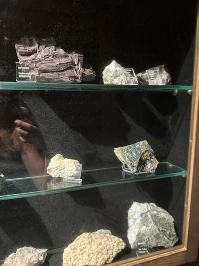This is a close-up, eye-level, medium shot of a display case at a museum in Schonach im Schwarzwald, Germany. The display case contains a collection of mineral specimens. The specimens are arranged on glass shelves.
The top shelf displays three mineral specimens. The largest specimen, on the left, is a dark red, layered rock labeled "ROTEISEN Flußspatgrube REINERZAU". To its right are two lighter colored crystalline specimens, each with a label. The left specimen is labeled "THAUMASIT PREHNIT Str. Vulkan HASLACH i.K.". The right specimen is labeled "PREHNIT CALCIT Str. Vulkan HASLACH i.K.".
The middle shelf has two specimens. On the left, a light-colored mineral is partially visible. On the right, a complex mineral specimen with green and blue hues sits on a small silver stand, labeled "AZURIT KUPFERERZ NEUBULACH". A reflection of a person's hand is visible on the glass of this shelf.
The bottom shelf displays two more mineral specimens. On the left, a light-colored crystalline specimen is visible. To its right, a larger, lighter gray specimen with darker inclusions is labeled "PEKTOLITH Str. Vulkan HASLACH i.K.".
The background of the display case is dark and textured. The sides of the display case are framed by dark wood.
