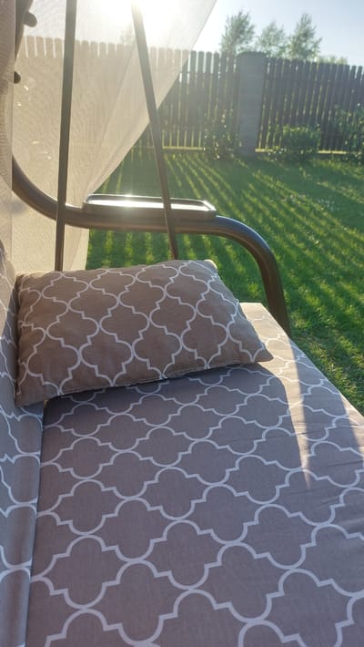 A close-up shot shows a cushion on a swing seat with a brown and white quatrefoil pattern. The cushion is placed on a larger seat cushion with the same pattern. A white mosquito net hangs over the seat, creating a soft, diffused light. In the background, a wooden fence encloses a green lawn, with trees visible behind the fence. The sun shines brightly, casting long shadows across the grass. The location is Masty, Belarus.