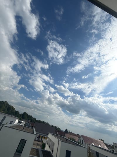 This is a landscape, vertically oriented, image taken on a sunny day in Biberach an der Riß, Germany. The photo captures a bright blue sky filled with a variety of white, fluffy clouds. The clouds are scattered across the sky, with some appearing more dense and textured than others.

In the lower right portion of the image, modern residential buildings are visible. These buildings have white facades, dark-framed windows, and flat or low-pitched roofs. Some of the roofs appear to be covered in gravel or decorative stones, while one is tiled in a reddish-brown color. The buildings are situated closely together, suggesting an urban or suburban setting.

A line of lush green trees runs along the horizon behind the buildings, providing a natural contrast to the architecture. The overall scene suggests a peaceful and pleasant day, likely in the late morning or early afternoon, given the bright sunlight and clear sky. There is no text visible in the image.