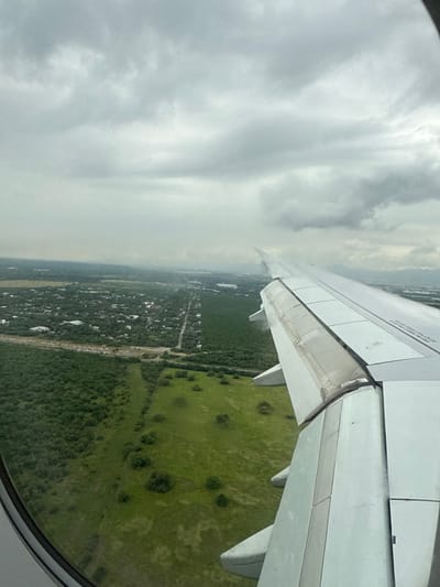 This aerial image, captured from the window of an aircraft, depicts a landscape within the city of Pesquería, Nuevo León, Mexico, under overcast skies. The right wing of the aircraft, with deployed flaps indicating descent or ascent, occupies the mid-right portion of the frame. The sky above is entirely covered by dense, grey clouds, suggesting low visibility or impending precipitation.
Below the aircraft, the terrain consists of a mixture of natural and developed areas. In the foreground and midground, large sections of dense green vegetation are visible, bordering expansive open grassy fields with scattered individual trees. A prominent straight road or dirt path runs horizontally across the midground, separating different types of land use. To the far left, a discernible residential area is present, characterized by numerous small buildings and a grid of roads, partially obscured by the distant haze. Further in the background, towards the center-right, indistinct structures or urbanized zones are visible through atmospheric haze, suggesting industrial or commercial development. Faint outlines of mountains rise on the distant right horizon. No individuals or specific actions beyond the aircraft's flight are discernible.