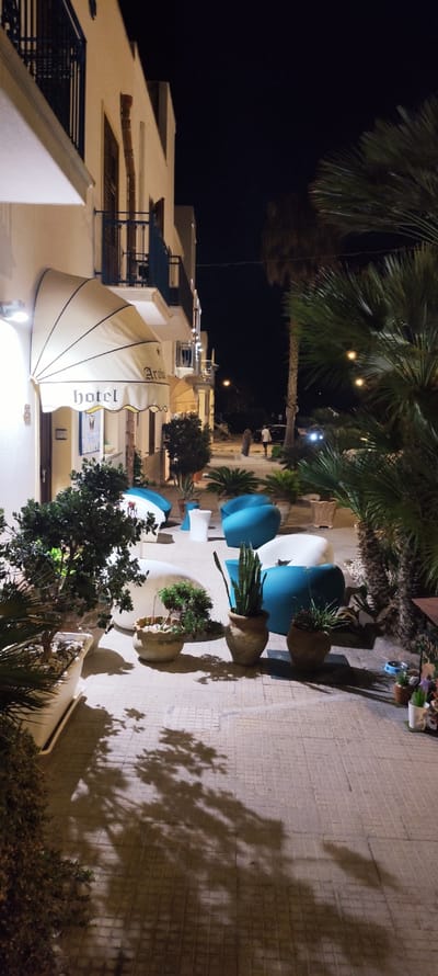 The image depicts the exterior of a hotel in San Vito Lo Capo, Italy, at night. A beige awning with "hotel" and "Araha" written on it is extended from the building. The building itself is light-colored with balconies. The ground level features a tiled walkway with several potted plants, including a large potted snake plant and a small bonsai. In the foreground, there are modern, irregularly shaped blue and white outdoor chairs. Shadows of trees are cast across the walkway. In the background, a few people are visible walking, and palm trees are present. The overall atmosphere is dimly lit, characteristic of nighttime.
