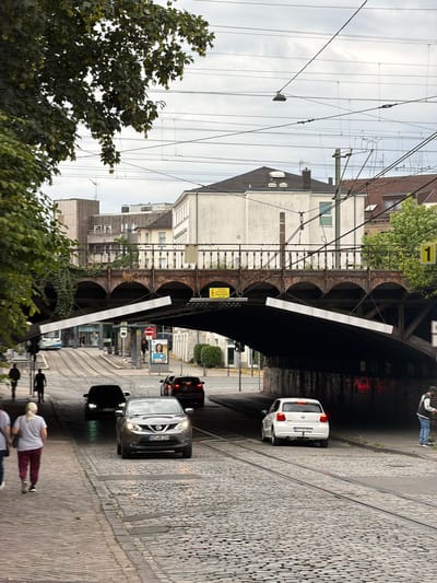 The image depicts a street scene in Witten, Germany, likely during daylight hours under an overcast sky. In the foreground, a cobblestone street runs towards a tunnel-like underpass beneath a raised railway bridge. The bridge is constructed with arched supports and has an ornate metal railing.
Several vehicles are present: a grey Nissan Qashqai is driving under the underpass, its headlights on, and a white Volkswagen Polo is visible exiting the underpass. Further back, a black car is approaching under the bridge, and a darker vehicle is partially obscured behind it. A tram is seen in the distance on the left, approaching the street.
On the left side of the street, two individuals are walking away from the camera, dressed casually. Further up the street, near the tram stop, several other figures are visible, including a person standing behind a red traffic sign and another person holding a poster with an image of a woman.
The environment includes a tree with green foliage on the left, obscuring part of the view, and buildings with windows and roofs visible behind the railway bridge. Overhead, a complex network of electrical wires, likely for the tram system, crisscrosses the sky. The overall atmosphere suggests an urban setting with active traffic and pedestrian movement.