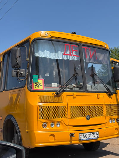 An eye-level, close-up, medium shot captures the front of a yellow school bus parked outdoors on a sunny day. The bus is the primary subject, with its large windshield prominently displaying the red Cyrillic word "ДЕТИ" (Children) in large letters. To the left side of the windshield, two green stickers with the year "2019" are visible, along with a yellow square sign depicting stylized figures of children running. A person in a striped shirt is partially visible through the windshield, sitting in the driver's seat. The bus features a large, mesh grille on the front, and the license plate, which reads "BY AC 3185-5", is located below the grille. The background consists of a clear blue sky and a few green trees on the right side, suggesting a rural or suburban setting. The overall scene appears to be in the Astrashitskagaradokski rural council, Belarus.