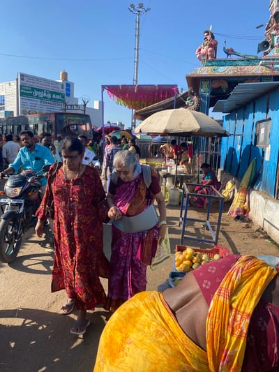 Two elderly women are walking on a dusty street in Chinnambedu, India. The woman on the left wears a long red and yellow patterned dress and grey sandals. She is holding a grey bag with a brown strap over her left shoulder. The woman on the right wears a purple sari and grey sandals. She is leaning on a grey support device around her waist. Both women are looking down as they walk.

In the background, a red bus is visible, along with several other people. A motorcycle is parked to the left of the women. A stall with fruit is set up to the right, with a woman in a pink sari sitting behind it. The street is lined with buildings and decorated with colorful banners. The sky is clear and blue.
