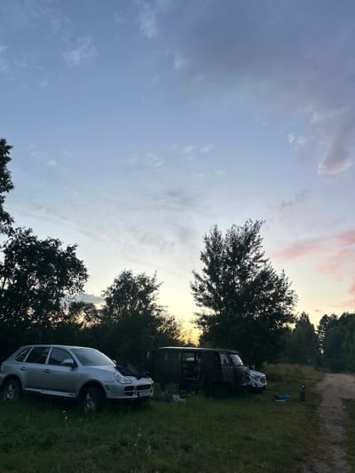The image depicts an outdoor scene at dusk, with a view of a rural landscape. In the foreground, on the left side of the frame, a silver SUV is parked on a grassy area. To the right of the SUV, a dark, military-style van is parked. In front of the van, a white car is partially visible. The vehicles appear to be stationary, and there are no people visibly interacting with them or in the immediate vicinity. The background is dominated by dense trees and foliage, with a dirt road curving into the distance on the right. The sky above is a gradient of blue and grey with wispy clouds, tinged with soft pink hues from the setting sun, suggesting the location is likely in a rural area, possibly near Тумілавіцкі сельскі Савет, Belarus, given the context. The overall impression is of a quiet, possibly remote, setting at the end of the day.