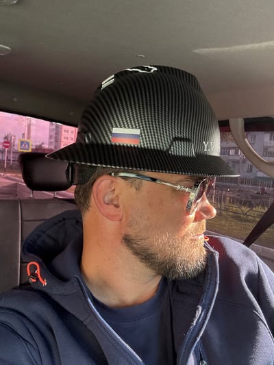 The image shows a man in a dark blue hoodie and a carbon fiber textured baseball cap with the Russian flag and the letters "Y.V.B." on it. He is wearing sunglasses and has a beard. The man is inside a vehicle, likely a car, as indicated by the headrest and interior of the car. Outside the car window, a city street is visible with apartment buildings and street signs. The lighting suggests it is daytime. The man is looking to his right, away from the camera. The overall scene is casual and appears to be taken in Usinsk, Russia, as suggested by the context.