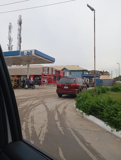 The image depicts an outdoor urban scene at the Mobil Filling Station located on 22 Road, Festac Town, Lagos, Nigeria. The foreground consists of a wet, light grey paved surface marked with numerous distinct tire tracks. Along the right edge, a low white-painted curb separates the pavement from a strip of dense green bushes.

In the mid-ground, a dark red minivan (Toyota Sienna, license plate MLN 105 OS) is visible from the rear, positioned on the right side of the paved area, facing away from the viewer. To the left, a Mobil gas station canopy, primarily blue and white with the "Mobil" logo, extends over fuel pumps. Directly adjacent to the Mobil station is a red KFC establishment, identifiable by its "KFC" signage and Colonel Sanders logo, including a "Drive-Thru" banner.

Several individuals are visible near the Mobil pumps; one person is positioned near the red minivan on the right. A yellow minibus is partially visible on the far left, near the gas pumps, and a motorcycle is parked nearby. The background features two tall telecommunications towers against an overcast sky. Further back, a blue building with a yellow roof is discernible behind the KFC, along with other indistinct urban structures. A utilitarian streetlamp pole stands vertically on the right side of the frame.