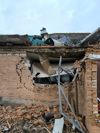 The image depicts a severely damaged brick building in Pysarivka, Ukraine, viewed from an exterior perspective under an overcast sky. The structure exhibits extensive destruction, with a large portion of its roof completely collapsed, revealing a chaotic tangle of broken wooden beams, corrugated metal, and torn insulation or fabric. A significant section of the upper brick wall below the roofline is missing, creating a large, irregular opening. The remaining red-orange brickwork shows multiple cracks and spallation. At the base of the wall, a considerable pile of debris consisting of fragmented bricks, splintered wood, twisted metal remnants, and other construction materials is accumulated on the ground. A bent, grey metallic support structure leans against the damaged wall, partially embedded in the rubble. No individuals are present in the scene.