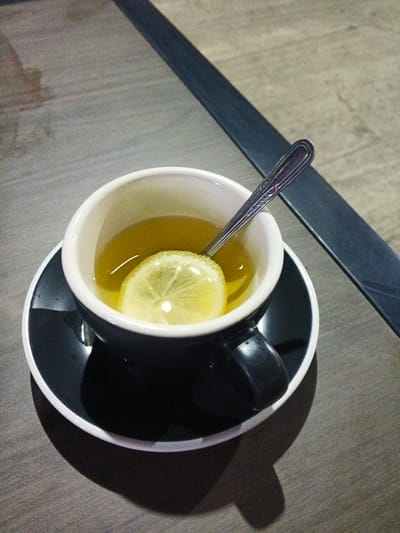 A black teacup with a white rim sits on a black saucer. The teacup is filled with a light-colored liquid, possibly tea, and a slice of lemon floats on top. A silver spoon rests in the cup, its handle extending towards the viewer. The cup and saucer are placed on a wooden table with a textured surface. In the background, a dark, raised surface runs diagonally across the frame. The lighting suggests an indoor setting, possibly a cafe or restaurant, with a soft glow illuminating the scene. The image is taken from a slightly elevated angle, looking down at the teacup. The overall mood is calm and inviting, with the warm tones of the tea and the clean lines of the tableware creating a sense of simple elegance. There is no visible text in the image.
