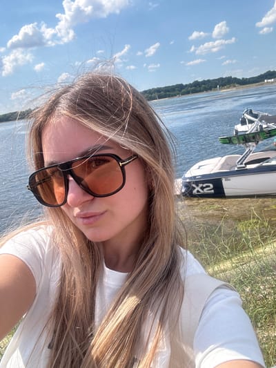 A young Caucasian woman with long blonde hair and sunglasses is taking a selfie near a body of water. The woman is wearing a white t-shirt and a light-colored vest. Behind her, a boat is docked near the shore, and there is a treeline in the distance under a blue sky with scattered clouds. The location is identified as Altenheim, Germany.