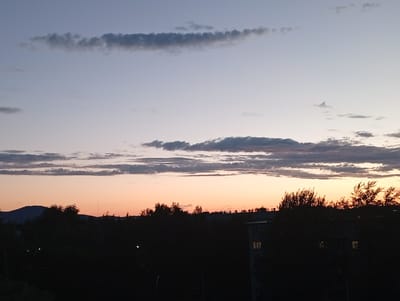 This media file depicts a twilight scene over Kyshtym, Russia. The sky transitions from a pale blue at the top to a warm peach and orange hue near the horizon, indicating either sunrise or sunset. A few wispy clouds are scattered across the sky, with some appearing as thin, elongated streaks and others as more textured formations.
Silhouetted against the colorful sky is a dark landscape featuring rolling hills or mountains in the distance to the left. In the foreground, the dense foliage of trees and the rooftops of buildings are visible as dark shapes. Several structures, likely apartment buildings, are discernible, some with illuminated windows suggesting interior lights are on. The overall impression is of a quiet evening or early morning in a town, with the natural beauty of the sky contrasting with the urban elements. There are no people or discernible objects in the foreground, and no visible text. The atmosphere conveyed is serene and peaceful.