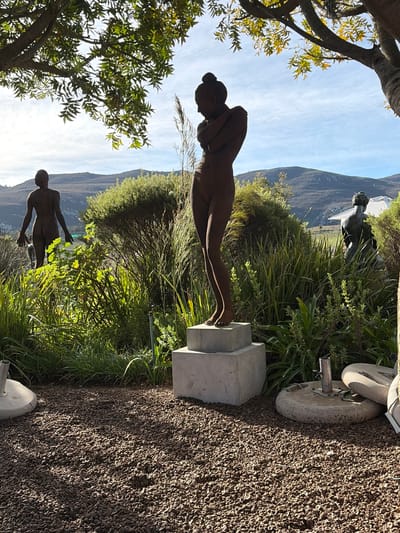 The image depicts an outdoor sculpture garden under a partly cloudy sky. The foreground is covered in dark brown gravel. In the mid-ground, several bronze or dark metal statues of nude human figures are visible. The most prominent statue is a female figure standing on a concrete pedestal, with her arms crossed over her chest. To the left, another nude female figure is seen from behind, walking away from the viewer. Further back and to the right, a third nude figure, possibly male, is depicted in a seated or crouching pose, partially obscured by vegetation. The background features lush green bushes and trees, with rolling hills and mountains in the distance, suggesting a rural or semi-rural setting. The presence of a large tree with green leaves in the upper left quadrant of the image indicates a natural environment. The lighting suggests it is daytime, possibly late morning or early afternoon, with sunlight casting shadows. The location is identified as Theewaterskloof Nu, South Africa.