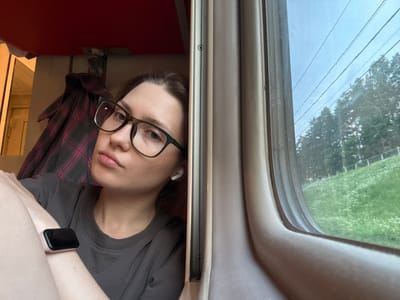 A young woman with glasses and brown hair, wearing a grey t-shirt and a smartwatch on her left wrist, is seen from the chest up, looking towards the camera. She is in a train compartment, with a red ceiling visible above and a plaid garment hanging on the wall behind her. To her right, a window shows a green, grassy landscape with trees and power lines outside, suggesting a rural or suburban setting. The scene's context is Povarovo, Russia, indicating the location of the train journey.