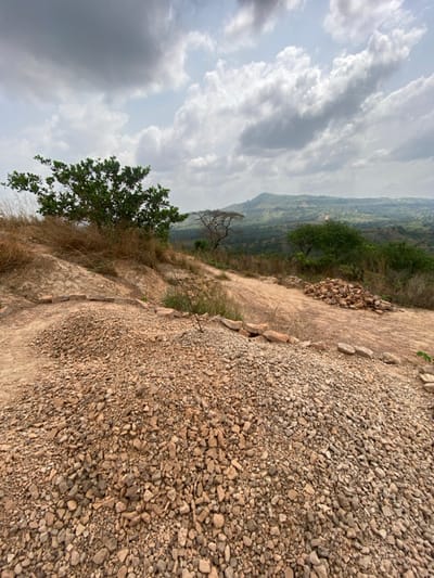 This image presents an elevated outdoor landscape scene in Ebuwana, Nigeria, under a sky with significant cumulus cloud cover.

In the foreground, large, irregular piles of light brown to reddish-brown gravel and fragmented rocks dominate the lower half of the frame, consistent with construction materials or excavated debris. A reddish-brown dirt path or disturbed ground is visible behind these piles, ascending slightly. Scattered individual rocks are present along the edges of the gravel mounds.

The midground features a dry, sloping terrain of reddish-brown earth interspersed with patches of dry, light-brown grass and sparse green vegetation. A dense, green-leafed tree stands on an elevated point on the left. Towards the center-right, a smaller tree with a gnarled, mostly bare trunk and branches is visible. Further right and slightly downhill, a smaller, structured pile of reddish-brown rocks is observed.

The background reveals a expansive green valley stretching into the distance, densely covered with trees and foliage. Multiple layers of undulating green hills or mountains extend towards the horizon, appearing slightly hazy.

No individuals or overt human actions are visible in the scene. The presence and arrangement of the gravel and rock piles strongly suggest prior or ongoing human activity related to construction, quarrying, or land development. Lighting is diffused, consistent with an overcast or partly cloudy day, resulting in soft or imperceptible shadows.