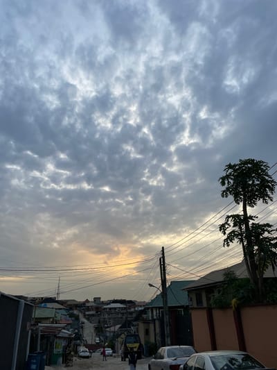 The image presents an overcast view in Isheri, Nigeria. The sky is predominantly filled with textured gray clouds. A slender tree with large, lobed leaves is visible in the upper left quadrant. Utility poles with numerous electrical wires crisscross the frame. Buildings, including residences and businesses, line a narrow street. Several parked vehicles are present, and a few individuals walk along the street. The scene appears to be a densely populated area with varied architectural styles.
