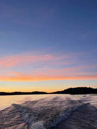 The image captures a sunset or sunrise view over a body of water within Ekerö kommun, Sweden, likely Lake Mälaren. The scene is dominated by a clear sky exhibiting a gradient from deep blue at the zenith, transitioning to violet, then a prominent horizontal band of vibrant pink and orange stratiform clouds, fading to a lighter yellow-orange at the horizon. Dark, forested landmasses, possibly islands or distant shorelines, form a continuous silhouette across the horizon line, separating the sky from the water. The water in the foreground and middle ground is dark, reflecting the sky's colors, with ripples and a distinct V-shaped wake from a moving vessel extending from the bottom center towards the middle distance. The wake features turbulent white foam at its crests, indicating forward motion away from the viewer. No people or specific identifiable objects other than the natural environment and boat wake are visible.
