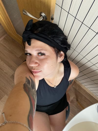The image shows a woman lying down, with her head resting on her arm. She has dark hair tied back with a black bandana and is wearing a black tank top. She has on makeup, including false eyelashes, and a silver necklace. There is a tattoo on her arm. The setting appears to be indoors, with a tiled wall and wooden floor visible. A door handle is also visible. The lighting suggests it is daytime. There is no visible text in the image.