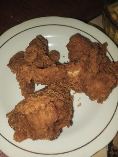 The scene is located in Doramamidi, India. The image presents a close-up, overhead view of a white, circular ceramic plate holding three pieces of fried chicken. Each chicken piece displays a dark golden-brown, visibly textured, crispy coating. The plate features a thin, brown or gold-colored band running along its outer edge. The background consists of a dark, likely wooden, surface in the upper left quadrant and a partially visible, blurred object with a yellow hue, possibly a bowl or dish, in the upper right. The lighting in the environment appears dim. No individuals or active interactions are depicted within the frame.