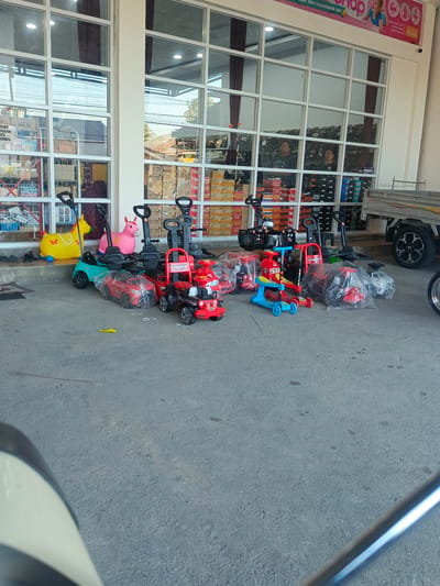 The image depicts a storefront in Tenggela, Indonesia, featuring a collection of children's ride-on toys displayed on a paved outdoor area. The storefront has large glass windows, through which various items are visible inside. In front of the store, several toys are arranged, including a yellow duck-shaped push car, a pink horse-shaped ride-on toy, a turquoise ride-on car, and several red and blue toy cars and scooters, some of which are partially covered in plastic. Behind the toys, a light-colored truck with a silver bed is parked. The ground is a textured gray asphalt. The overall scene suggests a retail environment for children's products, likely a toy store or a department store.