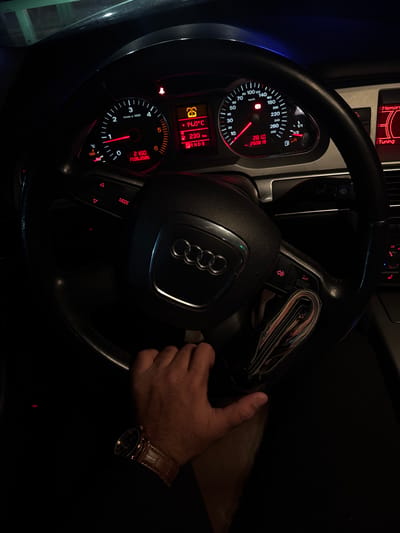 A dimly lit car interior is visible in the image. An Audi steering wheel dominates the scene, with the Audi logo prominently displayed in the center. The car's dashboard instruments are illuminated in red, displaying information like speed, RPM, time, date, and temperature. The time displayed is 21:50 on 17.05.2026. A man's hand wearing a brown leather watch is visible on the steering wheel, suggesting the driver's presence. A wallet, likely containing money, is positioned next to the steering wheel. The setting implies the car is stationary or being driven slowly in low light conditions. The location is not discernible from the image.