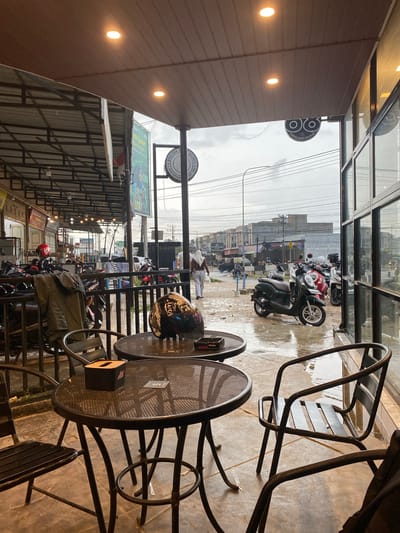 The media file captures a street scene in Keudeublang, Indonesia, viewed from inside a cafe or coffee shop. The image is taken through a large glass window, showing both the interior foreground and the exterior street.
In the cafe's foreground, several dark metal tables with mesh tops and chairs are visible. A black and white helmet rests on one table, next to a black tissue dispenser. Another table has a small sign that reads "MEJA 5". Overhead, bright ceiling lights are visible on what appears to be an awning or ceiling structure. A round sign displaying an "OO" logo and the text "COFFEE SHOP PREMIUM" is prominently featured.
Outside, the street surface is noticeably wet, indicating recent rainfall, and the sky is overcast, lending a slightly subdued atmosphere. Numerous motorcycles are parked along the street, a common sight in Indonesia. A few pedestrians are scattered along the road, including one person in light-colored clothing near the parked bikes. The street is lined with buildings, featuring various commercial establishments. Notable signs include a large green billboard bearing the text "UR Syariah", and a circular sign further down the street that reads "DOCTOR CLINIC" with "KEUDE GEUREUBA" below it, confirming the location. Overhead utility poles and a complex web of wires are visible against the cloudy sky. The overall scene depicts a typical, rainy day in an Indonesian town, observed from the comfort of a cafe. Other visible text includes "AMED", "ROY", "Jom", and "TIMUR SINITS".