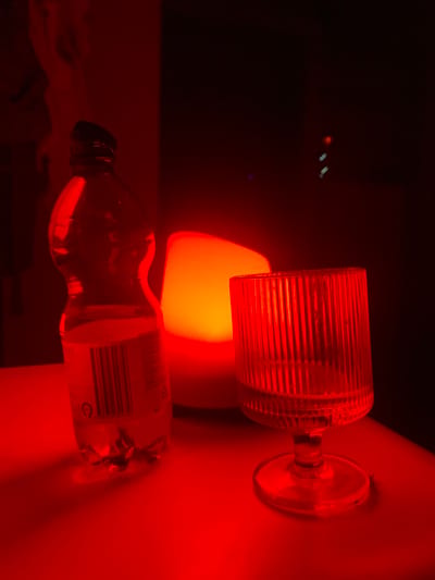 The image shows a close-up of a table with a bottle of water and a glass. The scene is illuminated by a red light, casting a strong red hue over everything. The bottle of water is on the left, with its label clearly visible, including a barcode. To the right of the bottle is a translucent orange lamp. Next to the lamp, on the right side of the image, is a glass with a fluted design, partially filled with a clear liquid. The background is dark, with some faint lights visible in the distance. The overall impression is one of a dimly lit, atmospheric setting. The image does not contain any information about the location in Loulé, Portugal.