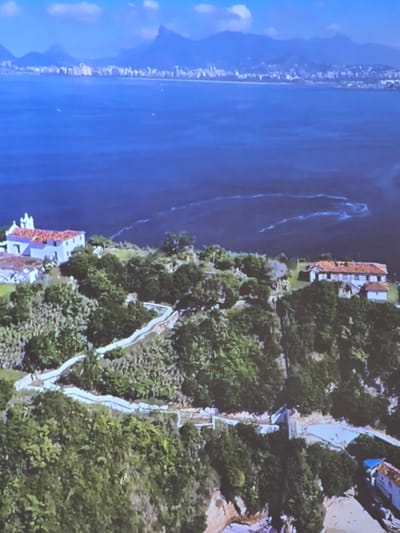 An aerial, wide-angle view captures a scenic coastal landscape. In the background, a sprawling city with white buildings and mountains rises from the horizon, met by a vast expanse of deep blue ocean. Wisps of white foam or clouds are visible on the water's surface. In the foreground, a verdant hillside is dotted with trees and a winding white path. Perched atop the hill are two distinct buildings: one a long, white structure with a red-tiled roof, and another smaller, white building with a red-tiled roof. The environment is lush with foliage, suggesting a tropical or subtropical climate. The scene is tranquil and picturesque, showcasing the juxtaposition of urban development, natural beauty, and architectural elements.