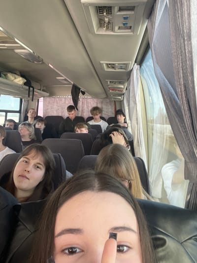 The image depicts the interior of a passenger bus, reportedly in transit within the city of Meshcheryaki, Russia. The perspective is a low-angle selfie, capturing approximately fifteen to twenty individuals, primarily young adults and teenagers, seated in rows.
In the immediate foreground, the upper portion of an individual's face is visible, including the eyes, forehead, and dark brown hair. A finger is shown adjusting a small, dark, rectangular object on the right eyebrow. Directly behind this individual, several passengers are seated. To the left, a young woman with brown hair, wearing a dark top, looks directly towards the camera. Behind her, an older woman with blonde hair and a dark top is partially visible. Further back, a young man in a dark hooded garment is seated prominently. Other young men and women are visible in various seats, some looking forward, others towards the camera, and some down. One passenger in the mid-background has visible earbuds.
The bus interior features dark grey fabric seats, light grey overhead compartments containing visible luggage items such as a white pillow-like object and a dark bag. The ceiling displays light fixtures, air vents, and passenger service units. The right side of the bus includes windows partially covered by patterned, light-colored curtains. The blurred view through the window indicates an outdoor environment consistent with open land or fields, suggesting the vehicle is in motion. No specific interactions between passengers are evident.