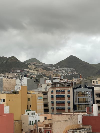 The image presents a panoramic view of a densely populated city nestled against a large, verdant mountain under an overcast sky. The scene is set in Santa Cruz de Tenerife, Spain, evident from the distinctive mountainous terrain and Mediterranean-style architecture.
The foreground and midground are dominated by numerous buildings of various heights and designs, mostly multi-story structures with flat roofs. Colors range from traditional whites, creams, and earthy tones like terracotta to more modern grey and black facades. Many windows feature blue or brown shutters and balconies. Rooftops display an array of satellite dishes, air conditioning units, and what appear to be solar panels. Electrical wires and poles are also visible throughout the urban sprawl. The city extends upwards, covering the lower slopes of the steep, green mountain, which forms a dramatic backdrop.
The weather is cloudy, with heavy grey clouds dominating the sky, suggesting a cool, possibly rainy day. The light is diffused, indicating daytime without direct sunlight. No people or specific activities are visible; the scene depicts a static urban and natural landscape.
A notable detail, visible in one of the provided crops, is a sign on a yellow building that reads: "100 años OPTICA RIEU". This indicates a long-standing optical business. The overall impression is one of a vibrant, compact city integrated into a striking natural environment.