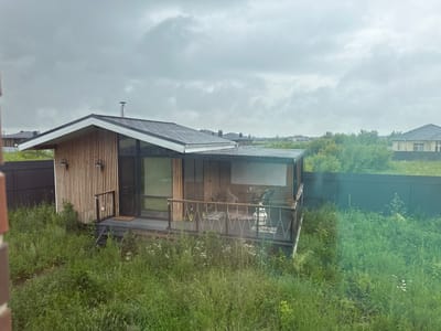 The image shows a modern, single-story wooden house with a dark gray pitched roof, located in the village of Kaipy, Russia. The house features vertical wooden siding and a large glass window. A dark gray deck with a decorative metal railing extends from the front of the house. On the deck, there is outdoor furniture, including a table and chairs, and a white decorative lantern. Steps lead from the deck down to the overgrown, grassy yard. The property is enclosed by a dark fence. In the background, other houses and trees are visible under a cloudy sky. The overall scene suggests a rural or suburban residential setting.
