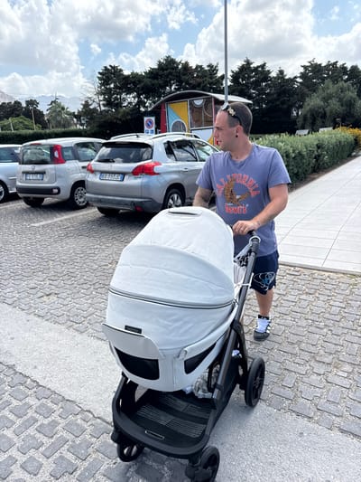 A man is pushing a stroller outdoors. He is wearing a blue t-shirt with a graphic of an eagle and "California Levi's" printed on it, and dark blue shorts. He has sunglasses on his head and is looking down at the stroller. The stroller is light gray and has a black frame. It is parked on a cobblestone surface next to a paved area. In the background, there are several cars parked in a row. The cars appear to be silver. One car has a license plate that reads "EX 353 JC". Another has "FE 370 LB". Behind the cars, there is a line of trees and a bright blue sky with white clouds. The scene suggests a sunny day. The man's body language, with his head down, could indicate focus or a slight weariness.