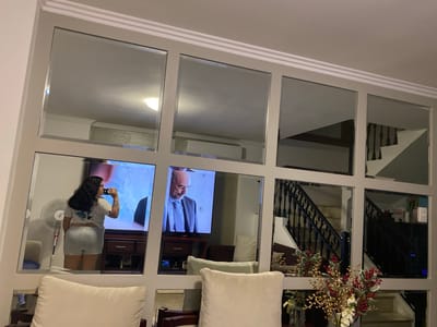A residential interior is depicted, featuring a wall adorned with a grid of square mirrors. The mirrors reflect various aspects of the room, including a woman drinking from a container, a television displaying a man in a suit, a staircase with black railings, furniture (including white cushions), and a floor fan. A floral arrangement sits in front of the mirror wall. It is not possible to confirm that the location is La Aurora, Ecuador with the given information.