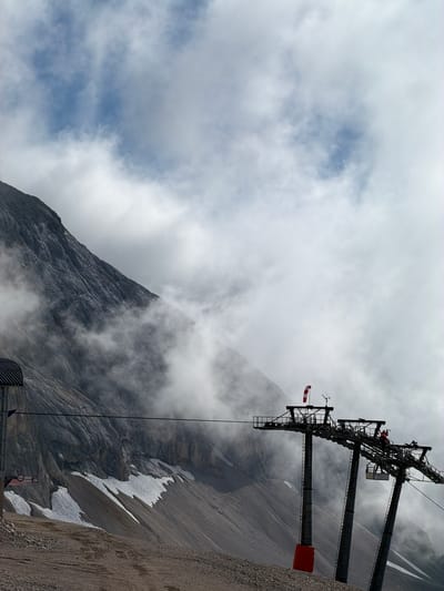 A ski lift station is shown in a mountainous, foggy environment. The ski lift station has three large support towers with a network of cables and pulleys for the ski lift. A windsock, red and white, is visible on top of one of the towers. The mountain face behind the ski lift is steep and rocky, with patches of snow. The sky is cloudy and overcast. The image appears to be taken from a lower elevation looking up towards the ski lift station and the mountain. This location is identified as Wildermieming, Austria.