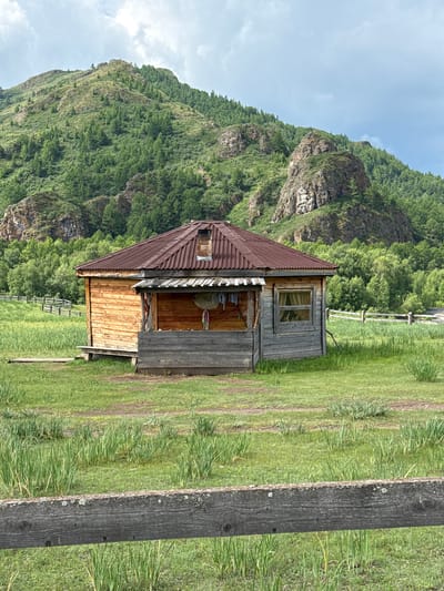 The image depicts a wooden cabin situated in a grassy field in Kazanovka, Russia. The cabin has a corrugated metal roof and is constructed from horizontal wooden planks. A porch area with a wooden railing is attached to the front. Behind the cabin, a lush green mountain covered in trees rises, with rocky outcrops visible. A wooden fence runs horizontally across the field in the middle ground, and another fence is partially visible in the foreground. The sky is partly cloudy. There are no people or animals visible in the scene.