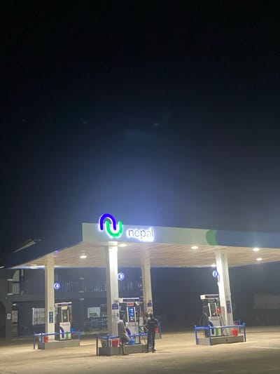 The image displays a nighttime view of a Nepal brand gas station located in Issele Uku, Nigeria. A brightly illuminated canopy, supported by multiple white pillars, features a prominent blue and green "Nepal" logo with white text. Recessed lights are active under the canopy, illuminating the service area. Two fuel dispenser islands are visible. At the leftmost island, two adult males are present. One individual, wearing a light-colored top and dark trousers, is standing near a fuel dispenser with several red jerry cans positioned on the paved ground. The second individual, dressed in dark clothing, is standing adjacent to the first, also in proximity to the fuel dispenser and containers. The actions indicate fuel is being dispensed into the portable containers. To the left of the gas station structure, a multi-story building with balconies and railings is visible, also under illumination. The surrounding environment is dark, consistent with nighttime conditions.