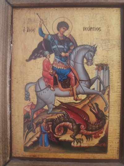 This is a religious icon depicting Saint George slaying a dragon. The central figure, Saint George, is shown mounted on a grey horse, wearing armor and a purple cloak. He is holding a spear, poised to strike the dragon. A smaller figure, possibly a princess or a maiden, is depicted below on the horse's rump, holding a jug. The dragon, a fearsome creature with red wings and sharp teeth, is on the ground, being attacked by Saint George. Another figure, a woman in blue, is shown to the left of the dragon, appearing to be an offering or a supplicant.
The scene is set against a gold-leaf background, typical of Orthodox icons, which signifies a sacred or heavenly space. The landscape is stylized, with rocky formations and a few trees. In the background, a white structure resembling a castle or tower with figures on top is visible.
The icon features Greek text. Above Saint George's head on the left are the words "Ο ΑΓΙΟΣ" (O Agios), meaning "The Saint". On the right, above the horse's head, is the name "ΓΕΩΡΓΙΟΣ" (Georgios), referring to Saint George.
The overall mood of the icon is one of triumph of good over evil, a common theme in religious art. There are no clear indications of time of day or weather, as the setting is symbolic. The location context of Neapoli Municipal Unit, Greece, is noted but not visually represented in the icon itself. The icon is framed by a simple wooden border.
