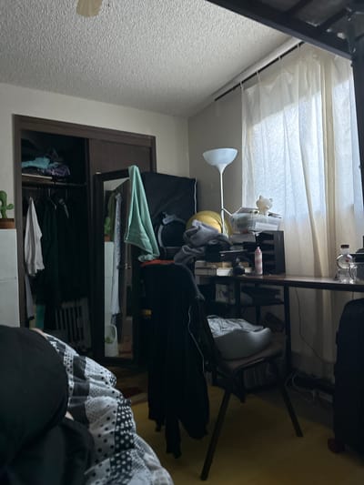 The image depicts a cluttered bedroom. On the left, a closet is partially open, revealing clothes on hangers and shelves with items. A small green cactus figurine sits on the top shelf. To the right of the closet, a desk is positioned against a wall, covered with various objects including a lamp, a plastic bin filled with items, and a pink bottle. A chair with a white cushion is placed in front of the desk. A large window covered by sheer white curtains occupies the background. The floor is a yellow linoleum. In the foreground, a person is lying in bed, covered by a black and white patterned comforter. The overall impression is of a lived-in and somewhat disorganized personal space. The location is not identifiable as Vancouver, United States of America.