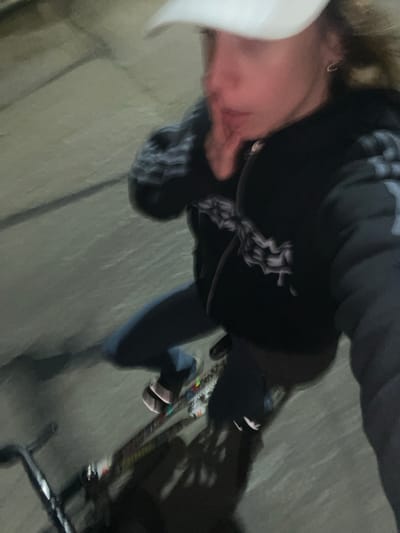 A blurry image shows a person wearing a white cap, dark hooded sweatshirt with a graphic design on the chest, and jeans. They are standing next to a bicycle on what appears to be a paved surface. The location is possibly Olivos, Argentina.
