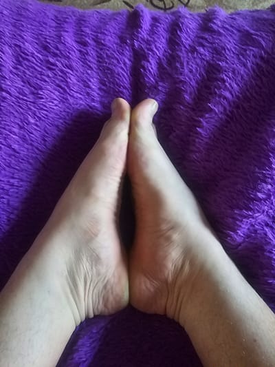Two bare feet are positioned close together, soles facing upwards, against a textured, vibrant purple surface. The feet are pale, with visible skin creases and some light hair on the ankles and lower legs. The purple surface appears soft and possibly furry, with a dense, short pile. The image is taken from a high angle, looking down at the feet. The location within the city of Таёжное, Russia is not discernible from the visual information provided.