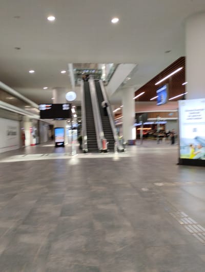 The image depicts the interior of a modern building, likely an airport terminal or a large shopping mall. The scene is characterized by its spaciousness and contemporary design. In the foreground, a gray tiled floor stretches out, showing a subtle pattern of light and dark tiles. Further back, an escalator is visible, ascending towards a higher level. People are present, though their forms are blurred due to motion, suggesting the photograph was taken while moving or with a slow shutter speed. One person is seen ascending the escalator, and other figures are scattered throughout the background, some appearing to be in shops or seating areas.

The environment features a high ceiling with recessed lighting. To the left, there are storefronts or kiosks. To the right, a large white advertisement panel stands prominently, featuring images and text, though the details are not entirely clear due to the blur. Above the escalator, a large digital display board with black background and white text is mounted. The overall lighting is bright and even, characteristic of indoor public spaces. The architectural elements suggest a new or well-maintained facility. There is no visual information in the image that specifically ties it to Tarsus, Turkey.