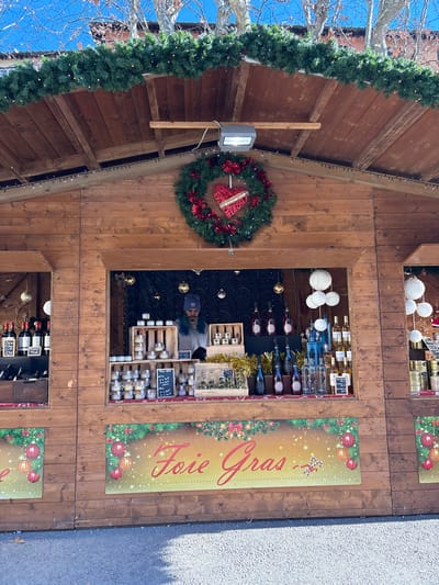 A wooden Christmas market stall, adorned with green garlands and a large wreath featuring a red heart, is depicted in the city of Lucca, Italy. A male vendor, wearing a blue beanie and a light-colored top, stands behind the counter within the open service window. The counter displays numerous small jars of pâté-like products, various bottles of wine and spirits, and multiple handwritten price tags. The stall's interior is decorated with golden baubles and white spherical lanterns, with a dark backdrop. A prominent banner along the lower front of the stall reads "Foie Gras" in red script, surrounded by festive ornaments. Above the stall, a clear blue sky is visible, along with bare tree branches and a section of a building with red roof tiles. The ground in front of the stall is a light grey, paved surface.