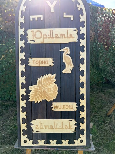 The image shows a wooden sign with carved text and images. The sign is dark brown with a decorative wooden border. There are four sections of text, each on a lighter wooden plank. The top plank has the Cyrillic letters "Юрлатмь". Below this, a wooden carving of a bird, possibly a heron or crane, is attached to the dark wood. The next plank reads "Торна" in Cyrillic. Below this is a carved image of a sprig of rowan berries. The final plank at the bottom reads "аlimalilak" in Latin script. There are small metallic studs holding the planks to the main sign. The sign is outdoors, with a camouflage net and foliage visible in the background, suggesting an outdoor event or market. The location is identified as Аргамак, Russia. The sign appears to be free-standing, mounted on a wooden easel. The lighting suggests it is daytime.