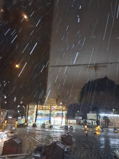 This image depicts an urban environment in ქალაქი ლანჩხუთი, Georgia, captured at night during active snowfall. The perspective is elevated, overlooking a city street or square. Numerous bright, elongated streaks of light indicative of falling snow are visible across the frame. The ground surfaces are dark, wet, and highly reflective, showing accumulated snow, slush, and tire tracks.
In the immediate foreground, two dark-roofed, small structures, resembling kiosks or temporary shelters, are partially visible. To the lower left, a large, illuminated number "26" decoration rests on the ground. Multiple vehicles are parked along the street and in designated areas, reflecting the ambient light. Several indistinct human figures are discernible walking on the wet surfaces in the mid-ground.
Across the street, a prominent multi-story commercial building dominates the background. Its ground floor features multiple storefronts with brightly illuminated interiors and signs. Upper floors also display illuminated windows. Decorative string lights are visible on the building's facade and potentially on nearby trees or poles. To the right of this central building, other structures, some with illuminated windows, and a few palm trees adorned with festive lights, are present. Overhead streetlights are visible as blurred bright spots in the upper left portion of the image.
